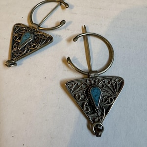 Puede incluir: Dos broches triangulares de filigrana plateada con piedras turquesa. Cada broche tiene un anillo circular con un pasador.