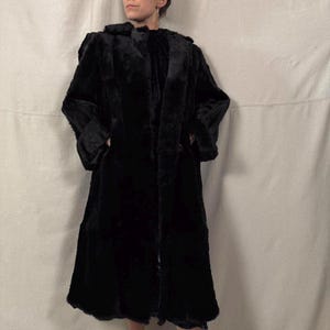 May include: A long, black faux fur coat with a wide collar and a button closure. The coat has a knee-length hemline and long sleeves. The coat is made of a soft, plush material and appears to be vintage.