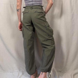 Può includere: Pantaloni cargo verde oliva con più tasche e coulisse alle caviglie. I pantaloni hanno una vita alta e una vestibilità rilassata. Il tessuto sembra essere un materiale resistente e tessuto.
