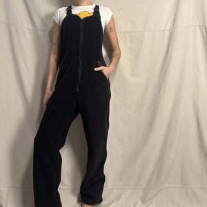 Puede incluir: Pantalón de peto de forro polar negro con cremallera en la parte delantera y detalles de costuras rojas. El pantalón de peto tiene dos bolsillos grandes en la parte delantera.