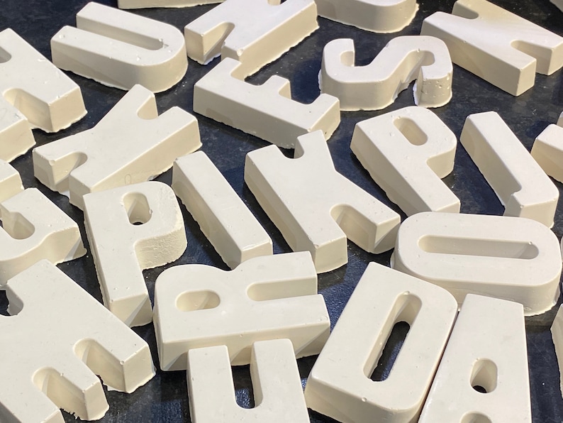 May include: White painted wooden letters of the alphabet, scattered on a black surface. The letters are all uppercase and are in various sizes.