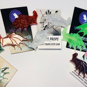 May include: A collection of bat wing-shaped accessories in various colors, including glittery orange, iridescent white, and green. The wings are displayed on cards with the text "Pixies Props" and social media handles.