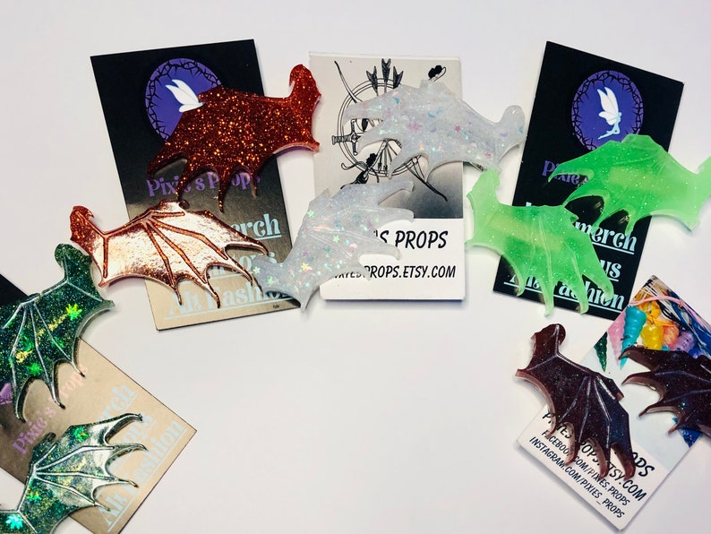 May include: A collection of bat wing-shaped accessories in various colors, including glittery orange, iridescent white, and green. The wings are displayed on cards with the text "Pixies Props" and social media handles.