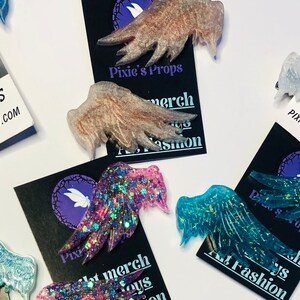 May include: Assortment of glittery resin fairy wings in various colors, including pink, blue, and clear. Each wing is detailed with sparkling glitter and rests on a black card with the text "Pixie's Props" and a website address.