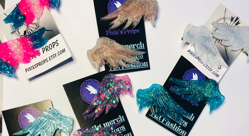 May include: Assortment of glittery resin fairy wings in various colors, including pink, blue, and clear. Each wing is detailed with sparkling glitter and rests on a black card with the text "Pixie's Props" and a website address.