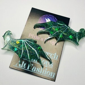 May include: Two glittery green dragon wing-shaped accessories. The wings have a detailed vein design and are decorated with sparkling glitter and small leaf-like shapes. The wings are on a card that says "Pixie's Props" and "Alt Fashion".