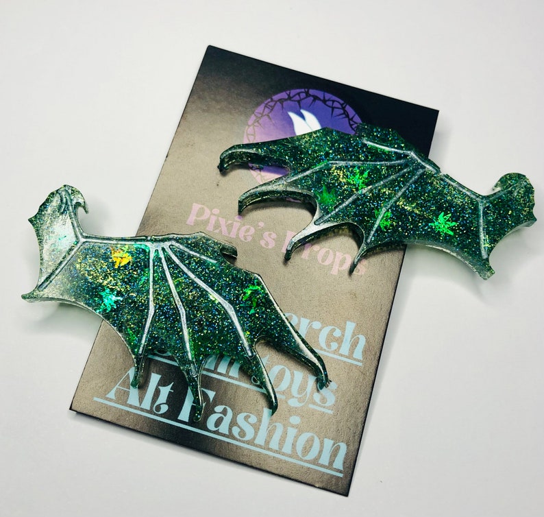 May include: Two glittery green dragon wing-shaped accessories. The wings have a detailed vein design and are decorated with sparkling glitter and small leaf-like shapes. The wings are on a card that says "Pixie's Props" and "Alt Fashion".