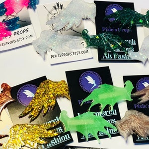May include: A collection of glittery wing-shaped accessories in various colors, including pink, blue, green, gold, and brown. Some wings are on cards with the text "Pixie's Props" and "Alt Fashion".