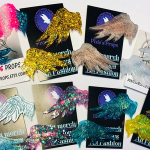 May include: Assorted glittery fairy wing accessories in various colors, including pink, blue, gold, and silver. Each wing is attached to a black card with the text "Pixie's Props" and "Art Fashion". The wings are designed for cosplay or costume use.