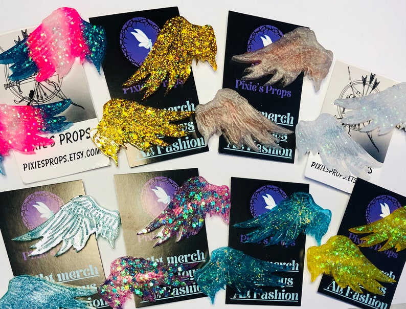 May include: Assorted glittery fairy wing accessories in various colors, including pink, blue, gold, and silver. Each wing is attached to a black card with the text "Pixie's Props" and "Art Fashion". The wings are designed for cosplay or costume use.