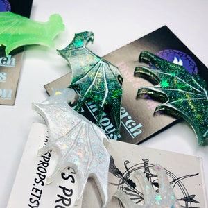 May include: Close-up of several glittery resin dragon wing-shaped ornaments in various colors, including green, white, and iridescent. Some wings are resting on cards with the text "Pixie's Porch" and "Pixie's Props.Etsy.com".
