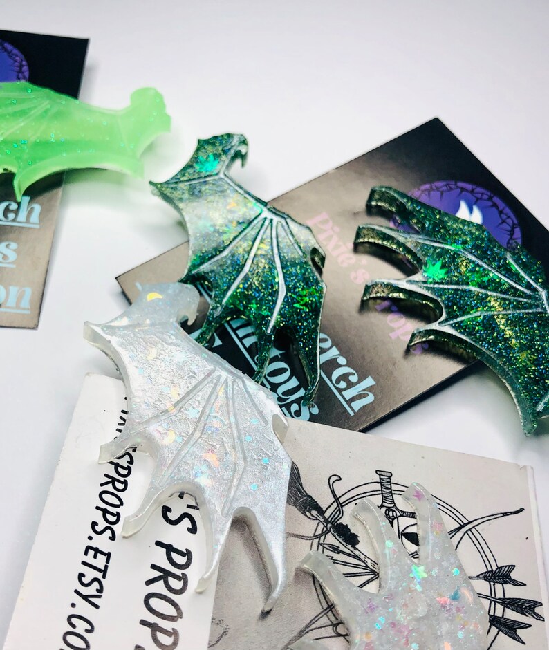 May include: Close-up of several glittery resin dragon wing-shaped ornaments in various colors, including green, white, and iridescent. Some wings are resting on cards with the text "Pixie's Porch" and "Pixie's Props.Etsy.com".