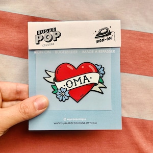 May include: Iron-on patch with a red heart and a banner that says "OMA" in a vintage tattoo style. The heart has a blue flower and green leaves on the side.
