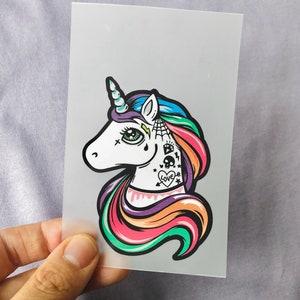 Könnte beinhalten: Ein farbenfrohes Einhorn mit einer Regenbogenmähne und einem Horn. Das Einhorn hat ein weißes Gesicht mit schwarzen Augen und ein schwarzes Herz mit dem Wort "love" auf der Wange. Das Einhorn hat weitere schwarze Tattoos auf seinem Gesicht, darunter ein Spinnennetz, einen Totenkopf, einen Blitz und einen Diamanten.