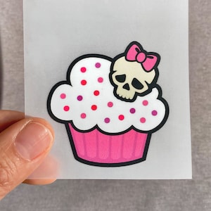 Könnte beinhalten: Ein rosa und weißer Cupcake mit rosa und roten Streuseln und einem weißen Totenkopf mit einer rosa Schleife oben drauf.