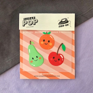 Puede incluir: Tres parches termoadhesivos con una pera, una naranja y una cereza con caras sonrientes. Los parches están sobre un fondo a rayas rosa y blanco con el texto "Sugar Pop Cologne" e "Iron-On" en negro.