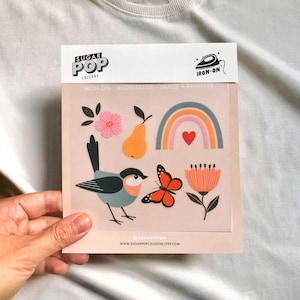 May include: A sheet of iron-on stickers featuring a rainbow, a pear, a bird, a butterfly, and flowers. The stickers are on a pink background and have a white border. The text "IRON-ON" is printed on the top of the sheet.