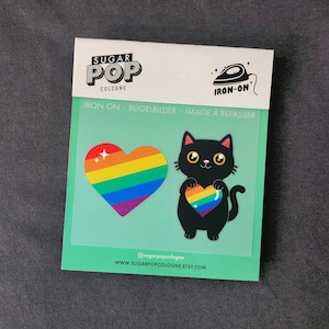 Könnte beinhalten: Zwei Bügelbilder, eines mit einem Regenbogenherz und das andere mit einer schwarzen Katze, die ein Regenbogenherz hält. Die Bügelbilder befinden sich auf einem weißen Hintergrund mit dem aufgedruckten Text "Sugar Pop Cologne" und "Iron-On".