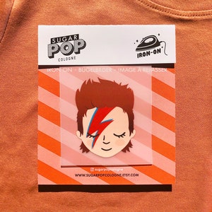 Könnte beinhalten: Bügeleisen-Patch mit einer Cartoon-Illustration von David Bowie mit einem roten Blitz auf seinem Gesicht. Der Patch befindet sich auf einem rosa-weiß gestreiften Hintergrund mit dem Text "Sugar Pop Cologne" und "Iron-on" auf der Verpackung.
