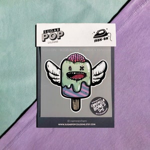 Könnte beinhalten: Bügeleisen-Aufnäher mit einem grünen und rosa Eis am Stiel mit Flügeln und einem Zombie-Gesicht. Der Aufnäher wurde von Cinderella Bones entworfen und trägt unten den Schriftzug "Sugar Pop Cologne".