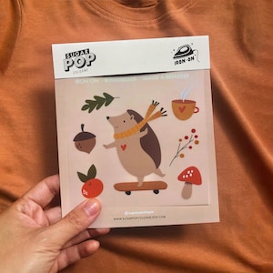 May include: Iron-on sticker sheet featuring a hedgehog on a skateboard, a cup of coffee, an acorn, a mushroom, and a tangerine. The sticker sheet is white with a pink background and the text "Iron-on - Bügelbilder - Image à repasser" at the top.