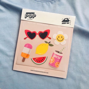 Op de afbeelding: Een vel met strijkbare stickers met een zomers thema. De stickers bevatten een paar hartvormige zonnebrillen, een glimlachende bloem, een ijslolly, een citroen, een watermeloenpunt en een blikje frisdrank met het woord "FRESH" erop. Het vel is gelabeld met "SUGAR POP COLOGNE" en "IRON-ON".
