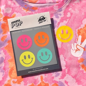 Puede incluir: Una hoja de parches termoadhesivos con cuatro caras sonrientes en diferentes colores: rosa, naranja, amarillo y verde. La hoja está etiquetada con "Sugar Pop Cologne" y "Iron-on - Bügelbilder - Image à repasser".