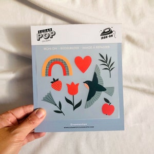 Puede incluir: Una hoja de pegatinas termoadhesivas con un arcoíris, un corazón, un tulipán, un pájaro, una fresa y una manzana. Las pegatinas están sobre un fondo azul y tienen el texto "IRON-ON" y "SUGAR POP COLOGNE" impreso en ellas.