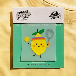 Könnte beinhalten: Bügeleisen-Aufnäher mit einer gelben Zitrone mit einem Regenbogen-Stirnband, die einen Tennisschläger und einen Tennisball hält. Der Aufnäher befindet sich auf einem grünen Hintergrund mit dem Text "SUGAR POP COLOGNE" und "IRON-ON" in Schwarz.