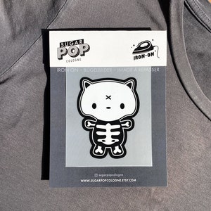 Peut inclure: Patch thermocollant noir et blanc représentant un chat de dessin animé avec un squelette à l'intérieur. Le patch est intitulé "Sugar Pop Cologne" et "Iron-On".