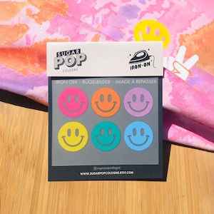 May include: A sheet of iron-on smiley face stickers in various colours. The sheet is labelled "Sugar Pop Cologne" and "Iron-On".