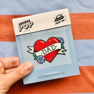 Könnte beinhalten: Bügeleisen-Aufnäher mit einem roten Herzen mit einem Banner, auf dem "DAD." steht. Das Herz ist von blauen Blumen und grünen Blättern umgeben. Der Aufnäher befindet sich auf einem weißen Hintergrund mit den aufgedruckten Worten "Iron-on" und "Bügelbilder".