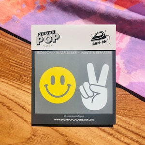 Könnte beinhalten: Bügelbild-Set mit einem gelben Smiley und einem weißen Peace-Zeichen. Das Bügelbild-Set ist auf einer grauen Karte verpackt, auf der der Text "Sugar Pop Cologne" und "Iron-on" aufgedruckt ist.