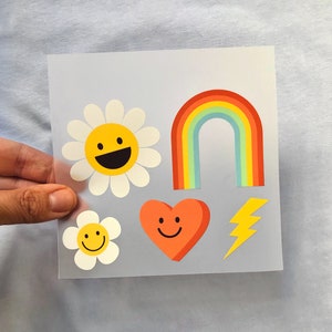 Puede incluir: Una hoja de pegatinas coloridas con un arcoíris, un corazón con una cara sonriente, una margarita con una cara sonriente y un rayo.