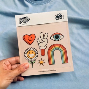 Puede incluir: Una hoja de parches termoadhesivos con fondo rosa. Los parches incluyen un arcoíris, un signo de paz, un corazón, una flor, una estrella y un ojo. La hoja está etiquetada con "Sugar Pop Cologne" y "Iron-On".