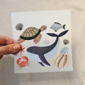 Può includere: Stampa quadrata con illustrazioni di creature marine. Il design include una tartaruga sorridente, una balena, un granchio, un pesce, una medusa, una stella marina e conchiglie nei toni del blu, marrone e arancione su sfondo bianco.