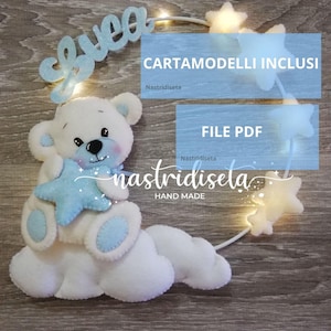 Può includere: Un orsacchiotto di feltro bianco fatto a mano che tiene una stella blu, appoggiato su una base a forma di nuvola. La parola "Sweet" è scritta in blu sopra l'orso. L'immagine include anche il testo "CARTAMODELLI INCLUSI" e "FILE PDF".