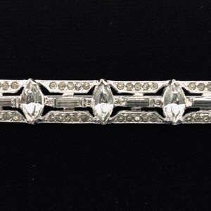 May include: A silver-toned bar brooch with a geometric design featuring five clear, faceted stones.
