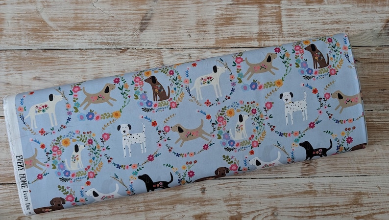 May include: A roll of light blue fabric with a repeating pattern of dogs and floral wreaths. The dogs are in brown, black, and white. The fabric has the text "EVERY HOME Every Dog" printed on the edge.
