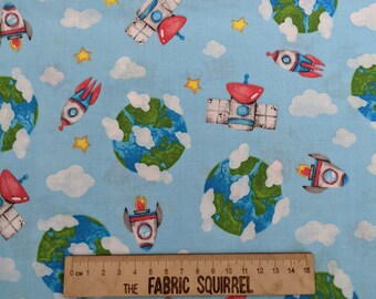 May include: A blue fabric with a repeating pattern of white clouds, green and blue planets, red and blue rockets, and yellow stars. The fabric has a ruler with the text "THE FABRIC SQUIRREL" printed on it.