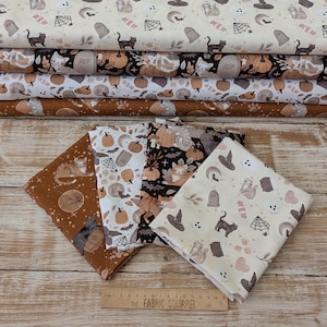 May include: Four folded pieces of cotton fabric featuring Halloween-themed patterns with cats, pumpkins, tombstones, and spiderwebs in shades of orange, black, and cream. A wooden ruler measuring 38 cm is placed at the bottom of the fabric pieces.