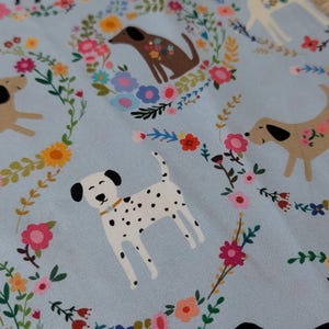 May include: Pale blue fabric with illustrations of dogs and floral wreaths. The dogs are in various breeds and colours, including black, brown, and white with black spots. The floral wreaths feature pink, yellow, and blue flowers, creating a cheerful design.