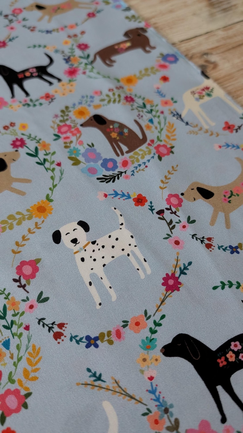 May include: Pale blue fabric with illustrations of dogs and floral wreaths. The dogs are in various breeds and colours, including black, brown, and white with black spots. The floral wreaths feature pink, yellow, and blue flowers, creating a cheerful design.