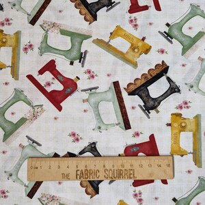 May include: A white fabric with a repeating pattern of vintage sewing machines in various colours, including red, green, yellow, and black. The fabric also features small pink roses scattered throughout. A wooden ruler with the text "THE FABRIC SQUIRREL" is visible at the bottom of the image.