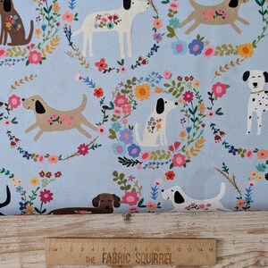 May include: Pale blue fabric featuring a repeating design of dogs and floral wreaths in multiple colours. The dogs are brown, white, and dalmatian-spotted. A wooden ruler with "THE FABRIC SQUIRREL" is at the bottom. Measurements are in centimetres.