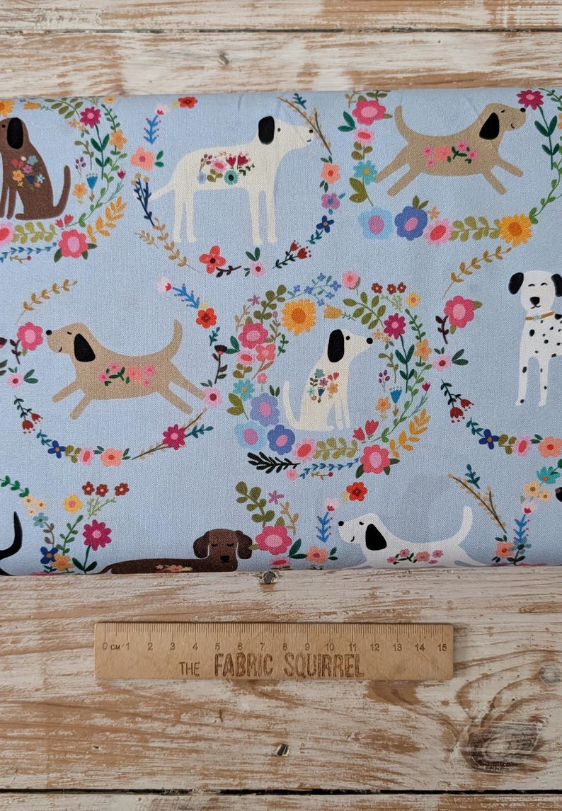 May include: Pale blue fabric featuring a repeating design of dogs and floral wreaths in multiple colours. The dogs are brown, white, and dalmatian-spotted. A wooden ruler with "THE FABRIC SQUIRREL" is at the bottom. Measurements are in centimetres.