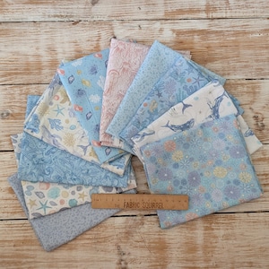 May include: A collection of folded fabric squares featuring various ocean-themed patterns in shades of blue, white, and soft pink. The prints include whales, seashells, starfish, and coral. A wooden ruler measuring 38 cm is placed across the fabric.