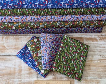 May include: A selection of fabric bolts showcasing nature-inspired patterns. Designs include mushrooms, leaves, and floral arrangements in blue, green, purple, and brown. The fabrics are displayed on a light wood surface.