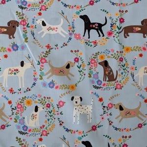 May include: Pale blue fabric featuring a repeating pattern of dogs and floral wreaths. The dogs are depicted in various colours, including white, brown, and black, with floral details. A cheerful and colourful design.
