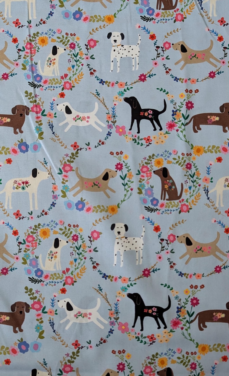 May include: Pale blue fabric featuring a repeating pattern of dogs and floral wreaths. The dogs are depicted in various colours, including white, brown, and black, with floral details. A cheerful and colourful design.